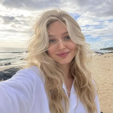 Blonde virtual influencer taking a beach selfie in a light lavender sweater, with ocean waves and cloudy sky behind her